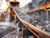 Close-up of rocks on a conveyor belt system in an industrial mining or quarry environment with heavy machinery in the background.